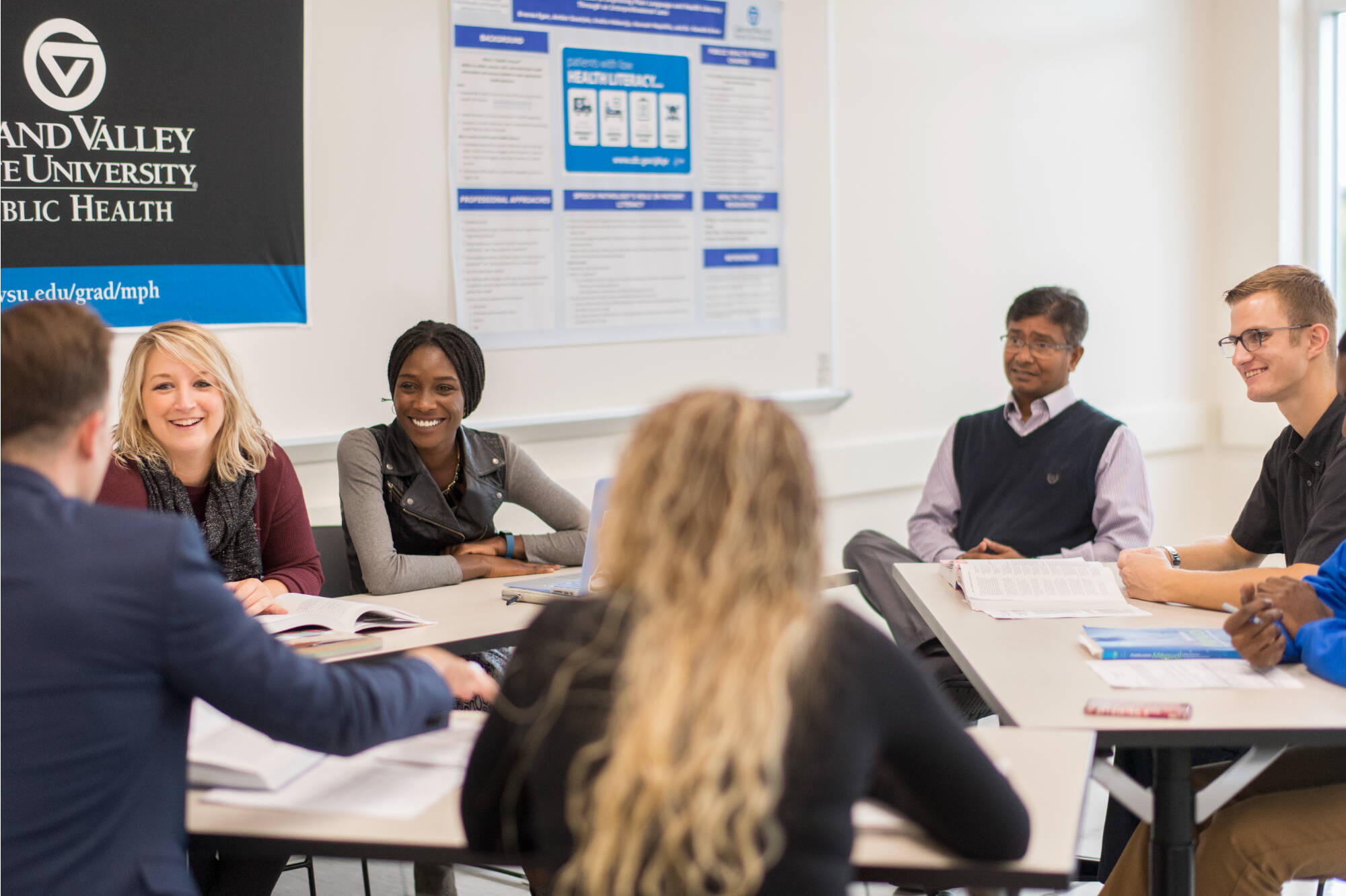 A group of students and faculty sit around a table in a classroom at Grand Valley State University. They are engaged in discussion, with papers and books spread out, and a banner on the wall displaying “Grand Valley State University School of Public...
